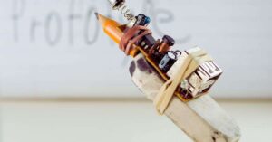 Close-up of a pencil attached to an eraser with rubber bands, fulfilling prototyping needs alongside a small electronic circuit and USB port. The background features blurred text on a whiteboard.