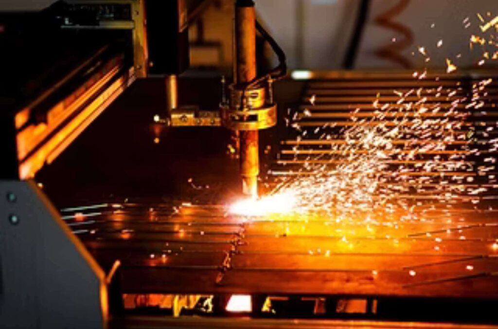 A CNC plasma cutting machine slices through a sheet of metal, creating bright orange sparks and a glowing cut line in an industrial setting.