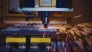 A CNC machining foundry machine cuts metal on a worktable, producing bright orange sparks that fly outward as the tool drills into the material in an industrial workshop.