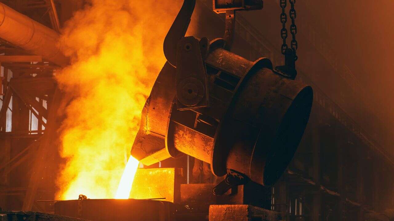 Molten metal pours from a large industrial ladle in a steel foundry, illuminating the dark factory with a bright orange glow. Machinery and metal structures form the backdrop of this heavy-duty casting process.