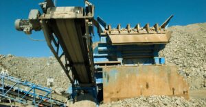 A large industrial rock crusher with a conveyor belt processes rocks at a quarry, though crusher output is declining, as seen by smaller piles of gravel and stones under a clear blue sky.