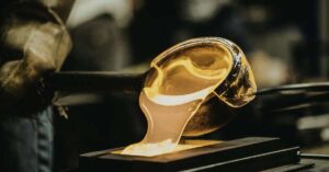A close-up of molten glass being poured from a ladle onto a surface during a glassblowing process. The glowing orange liquid, often assessed using casting inspection methods, contrasts with the dark, blurred background.